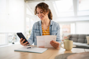 Young woman using smartphone while reviewing documents at home. Pay down your mortgage with small extra payments and prepayment strategies
